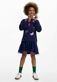 Donkerblauwe sweatshirt met geborduurde patches, gedragen over een navy tule rok. Gecombineerd met witte sneakers en groene sokken.