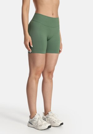 Kvinde iført olivengrønne højttaljede cykelshorts og hvide atletiske sneakers, stående mod en ensfarvet baggrund.