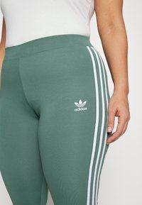 Gröna sportleggings i mjukt tyg, med vita sidostreck och en liten vit Adidas-logotyp nära midjan.