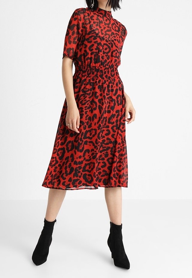 Femme portant une robe midi rouge à imprimé léopard noir, manches courtes, taille élastique, associée à des bottines noires.