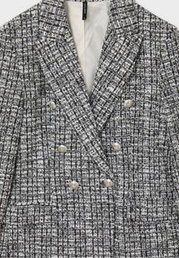Blazer croisé en tissu texturé noir et blanc à motif à carreaux. Des boutons argentés ornent le devant et les poches.