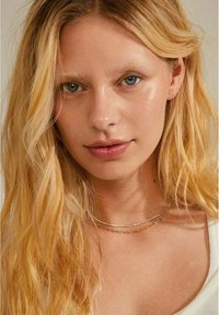 Des colliers superposés en or et en argent sur un teint de peau clair, featuring des chaînes délicates et une texture lisse et brillante. L'arrière-plan est neutre.