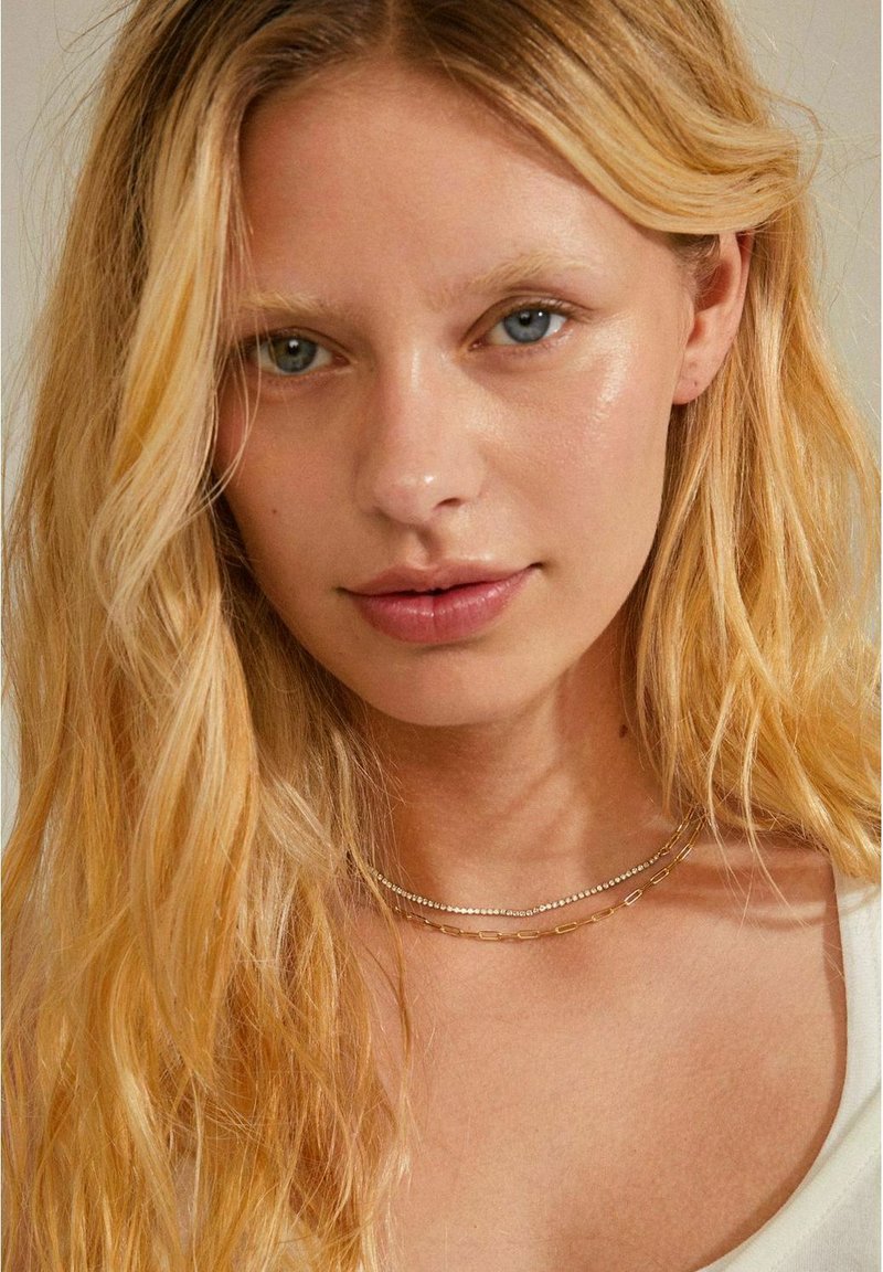 Des colliers superposés en or et en argent sur un teint de peau clair, featuring des chaînes délicates et une texture lisse et brillante. L'arrière-plan est neutre.