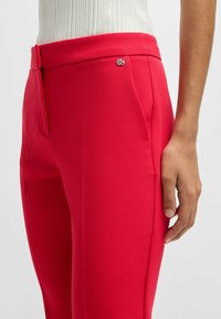 Pantaloni rossi attillati realizzati in tessuto liscio, dotati di passante per cintura e un accento metallico del logo sulla vita. Visibile il posizionamento delle mani sui fianchi.