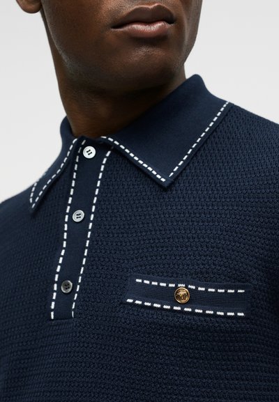 Gros plan d'une personne portant un polo texturé bleu marine avec une bordure blanche surpiquée, trois boutons et une petite poche avec un détail de bouton doré.