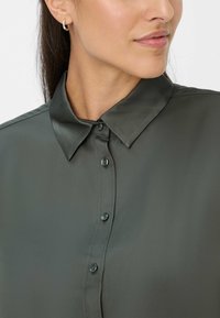 Blusa di seta verde scuro con colletto a punta, chiusura con bottoni e lieve lucentezza. Primo piano sul colletto e sui bottoni superiori.