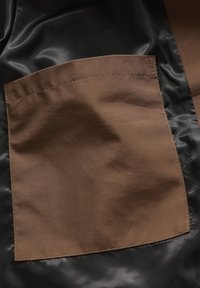 Tela de satén negra con un gran bolsillo parche marrón. El bolsillo tiene bordes cosidos, creando un contraste suave contra la textura brillante.