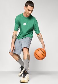 Grøn kortærmet skjorte med logo, grå træningsshorts, grå sokker med logo, sort og hvidt sneakers, og en basketball.