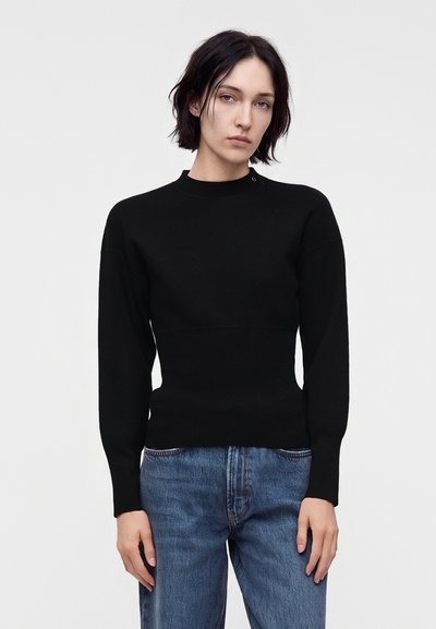Femme aux cheveux courts et foncés portant un pull noir moulant à manches longues et un jean bleu, debout devant un fond clair uni.