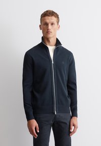 Mannensweater in marineblauw met ritssluiting in textuur breiwerk, voorzien van een hoge kraag en zilveren rits. Gecombineerd met donkere broek voor een samenhangende look.