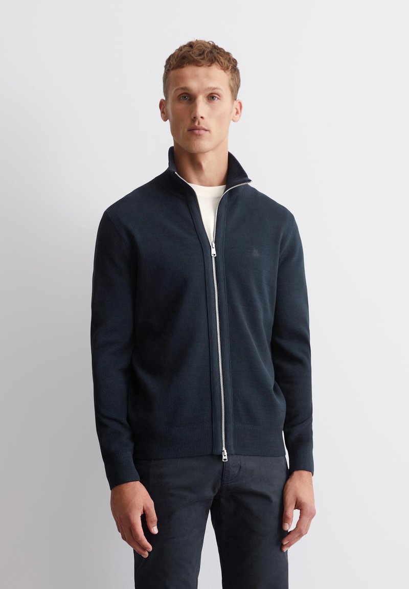 Mannensweater in marineblauw met ritssluiting in textuur breiwerk, voorzien van een hoge kraag en zilveren rits. Gecombineerd met donkere broek voor een samenhangende look.