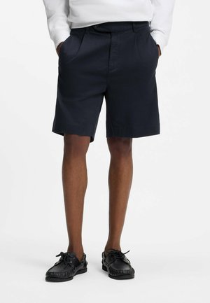 Person iført sorte shorts med sidelommer, en hvid langærmet top og sorte snøresko af mokasin-typen, stående med hænderne i lommerne.