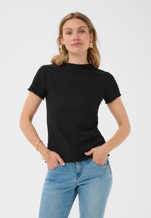 Vrouw die een zwart shirt met korte mouwen en lichtblauwe spijkerbroek draagt, met handen in de zakken, gouden creolen oorbellen en armbanden aan de linker pols.
