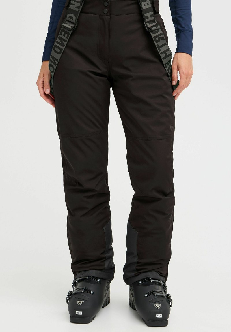 Black ski pants with adjustable straps, a smooth texture, button closure, and reinforced areas at the ankles, worn with black ski boots.