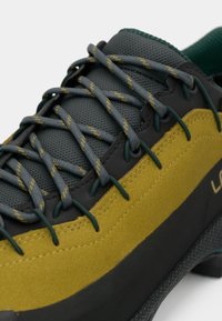 Avalynė su daugialype medžiagų konstrukcija, kuri apima pilką tinklinį audinį, geltoną zomšą ir juodus akcentus. Tekstūruotas paviršius ir akcentuota pilka raišteliais.