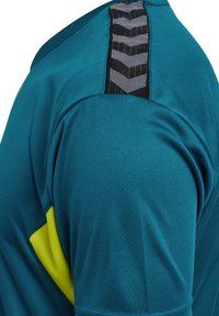 Nahaufnahme eines blau-grünen Sportshirt-Ärmels mit einem schwarzen Chevron-Musterstreifen und einem gelben dreieckigen Detail nahe der Achselhöhle.