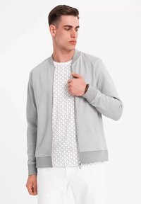 Bómber de color gris claro con cierre delantero, puños y dobladillo acanalados. Lleva encima una camisa blanca con un diseño geométrico.