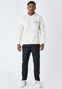 Felpa beige con cappuccio e coulisse, tasca frontale e grafica blu. Pantaloni cargo neri con più tasche. Sneakers bianche con dettagli neri.