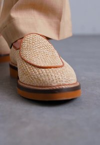 Mocassins tissés beiges avec des bordures en cuir marron, un bout rond et une semelle épaisse couleur tan, sur un fond gris.