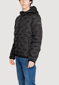 Giacca imbottita nera con cappuccio, dotata di zip frontale, polsini elastici e un patch con logo sulla manica sinistra. Materiale morbido e leggero.