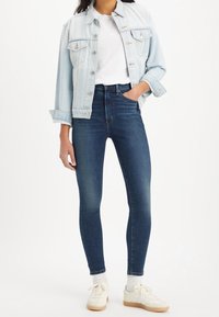 Chaqueta de mezclilla azul claro con dos bolsillos en el pecho sobre una camiseta blanca, combinada con jeans skinny azul oscuro y zapatillas blancas con suelas beige.