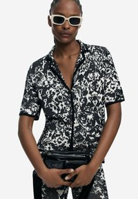 Black and white short-sleeve shirt featuring a floral pattern, button front, and black trim. Model holds a black multi-pocket bag.
