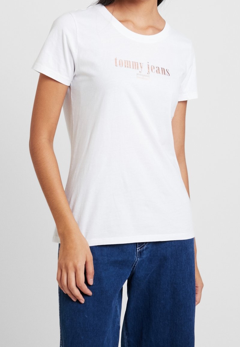 Žena v bílém tričku s krátkým rukávem, na kterém je nápis "tommy jeans" v světlé růžové barvě, a modrých džínech, stojící na jednobarevném pozadí.