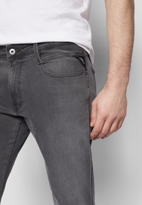 Homme portant un jean gris ajusté et une chemise blanche, la main droite détendue à côté de sa cuisse sur un fond uni.