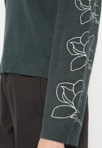 Pull en vert foncé avec un tricot texturé, présentant une broderie florale blanche le long des manches et un ourlet côtelé.