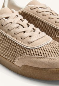 Beige színű sportcipő, hálós felsőrésszel, zamak díszítéssel, kerek fűzőkkel és gumitalppal, amely texturált mintázatot és sima felületet mutat.