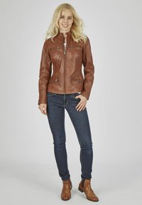 Braune Lederjacke mit einer taillierten Passform und hohem Kragen, ausgestattet mit zwei Reißverschlussfronttaschen und einer glatten Textur, kombiniert mit dunklen Jeans und Stiefeletten.