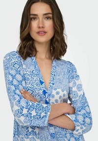 Blusa a fiori blu e bianca con scollo a V, maniche lunghe e dettagli con bottoni, caratterizzata da design intricati e una consistenza di tessuto liscia.