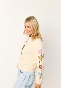 Ξανθιά γυναίκα που χαμογελά, φορόντας εκρού ζακέτα με πολύχρωμη ανθική και αφηρημένη κεντητική στο μανίκι, συνδυασμένη με ανοιχτό μπλε τζιν, πάνω σε λευκό φόντο.