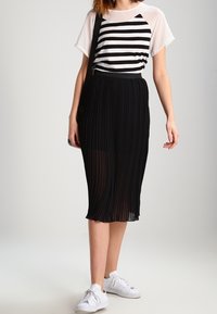 Svart plisserad midi kjol i kombination med en vit och svart randig kortärmad topp och vita sportskor, samt en axelväska.