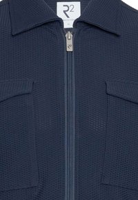 Marineblauwe zip-up jack met een gestructureerde stof, twee voorzakken en een puntkraag. Voorzien van een metalen rits en een label met de tekst "R2 Amsterdam, XL."