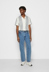Camicia a maniche corte grigia chiara e bianca, con due tasche sul petto. Indossata con jeans azzurri e sneakers bianche, in piedi di fronte a uno sfondo semplice.