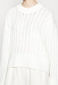 Weißer Strickpullover mit geripptem Muster, Bootsausschnitt und langen Ärmeln. Weiches Material und lockere Passform. Verfügt über eine leicht verkürzte Silhouette.