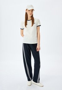 Hvid t-shirt med marineblå detaljer og logo, parret med marineblå bukser med hvide side-striber, samt en hvid kasket. Fodtøj er hvide sneakers.