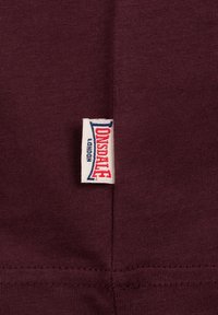 Burgundi színű pamut anyag, látható varrással és egy fehér címkével, amelyen vörös és kék betűkkel a "Lonsdale London" felirat található az oldalon.
