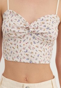 Top court floral avec un design de nœud à l'avant ; fabriqué en tissu texturé présentant de petites fleurs roses, jaunes et violettes sur un fond crème.