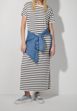 Personne portant une robe maxi rayée noire et blanche avec une veste en jean bleue nouée autour de la taille et des baskets bleu clair.