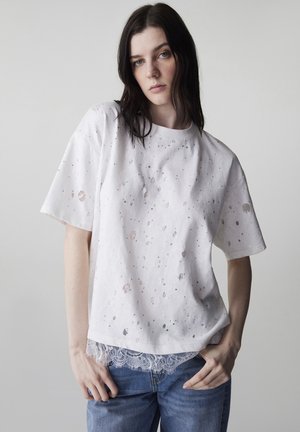 Mujer con cabello oscuro vestida con una camiseta blanca de gran tamaño con pequeños agujeros y ribete de encaje, combinada con jeans azules, sobre un fondo liso.