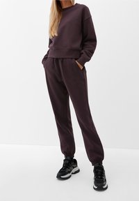 Felpa in misto cotone di colore viola scuro con scollo tondo, abbinata a pantaloni jogger coordinati con polsini elasticizzati e tasche. Include sneakers nere.