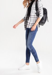 Jeans blu slim fit, camicia a quadri grigia e bianca con bottoni, canotta nera, sneakers bianche e zaino nero.