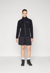 Giacca in pile nera con zip e colletto alto. Abbinata a pantaloni cargo neri e stivali da trekking neri. Calzini bianchi visibili sopra gli stivali.