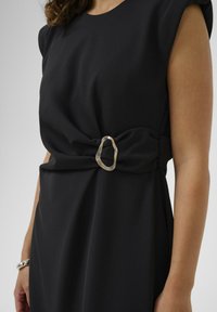Crna haljina s kratkim rukavima, s naglašenim okrenutim pojasom koji krasi nerazmjerni metalni kopč. Glatka tekstura tkanine.