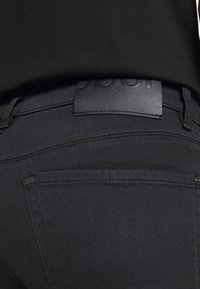 Vaqueros de mezclilla negra con una textura suave, con una etiqueta de cuero en la cintura y detalles sutiles de costura. La vista trasera destaca el diseño de los bolsillos.