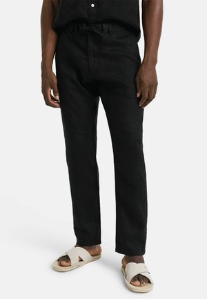 Mann trägt schwarze Hose und beige Sandalen mit gekreuzten Riemen, steht vor einem schlichten weißen Hintergrund.
