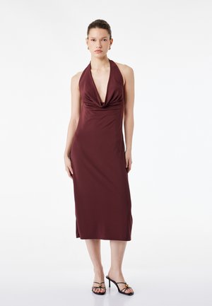 Femme debout portant une robe midi bordeaux sans manches avec un col drapé et des sandales à talons ouvertes noires sur fond blanc.