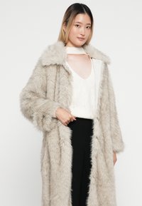 Cappotto lungo e morbido beige con colletto ampio e maniche piene, abbinato a un maglione bianco strutturato e pantaloni neri. Tessuto liscio e soffice.